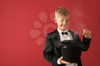 Cute little magician showing trick with hat on color background�