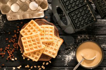 Wooden board with tasty waffles, maker machine and bowl with batter on dark wooden table�