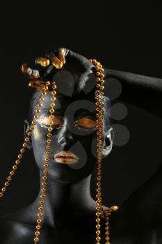 Beautiful woman with black and golden paint on her body holding beads against dark background�