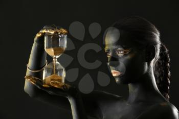 Beautiful woman with black and golden paint on her body holding hourglass against dark background�