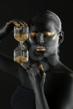 Beautiful woman with black and golden paint on her body holding hourglass against dark background�