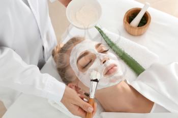 Cosmetologist applying mask with aloe vera extract onto face of young woman in beauty salon�