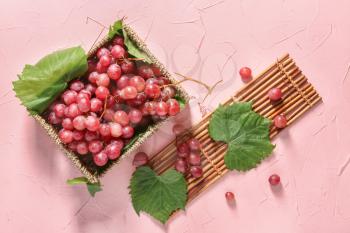 Wicker box with ripe grapes and bamboo mat on color background�