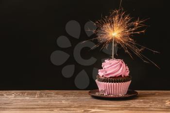 Delicious birthday cupcake with firework candle on table against dark background 