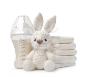 Baby bottle with milk, bunny toy and stack of diapers on white background�