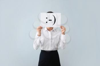 Young woman hiding face behind sheet of paper with drawn emoticon on light background�
