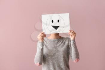 Young woman hiding face behind sheet of paper with drawn emoticon on color background�