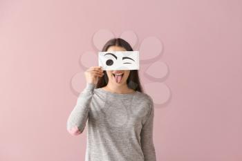 Emotional young woman hiding face behind sheet of paper with drawn eyes on color background�