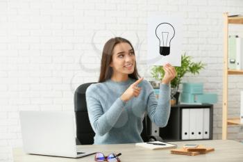 Emotional young woman holding paper sheet with drawn light bulb as symbol of idea while sitting at table�