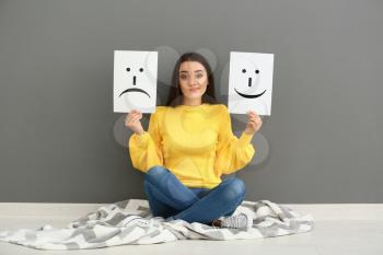 Emotional woman holding sheets of paper with drawn emoticons while sitting near grey wall�