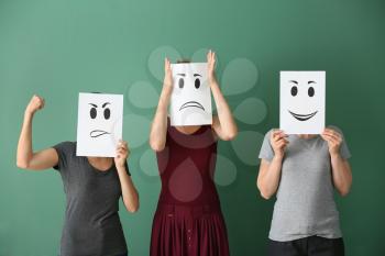 Young women hiding faces behind sheets of paper with drawn emoticons on color background�