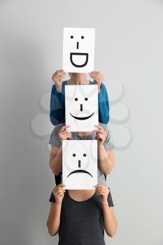 Young women hiding faces behind sheets of paper with drawn emoticons on light background�