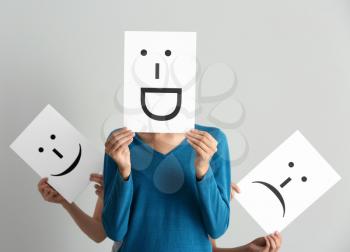Young women hiding faces behind sheets of paper with drawn emoticons on light background�