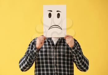 Man hiding face behind sheet of paper with drawn emoticon on color background�