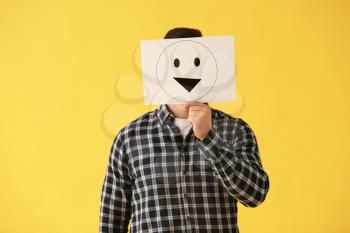 Man hiding face behind sheet of paper with drawn emoticon on color background�