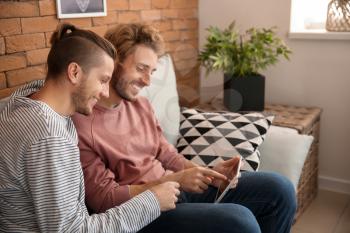 Happy young gay couple with tablet PC sitting on sofa at home�