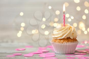 Delicious birthday cupcake with burning candle on grey table  against blurred lights�
