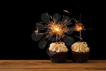 Tasty chocolate cupcakes with sparkler on wooden table against dark background�