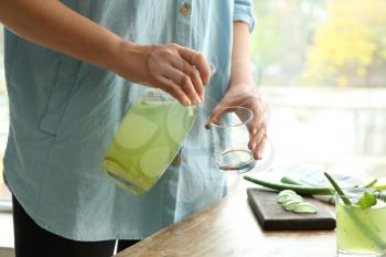 Woman pouring aloe vera cocktail from jug into glass at wooden table�