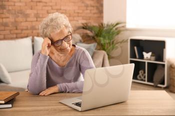 Senior woman suffering from headache while sitting at table with laptop�