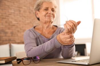 Senior woman suffering from pain in wrist while sitting at table with laptop�