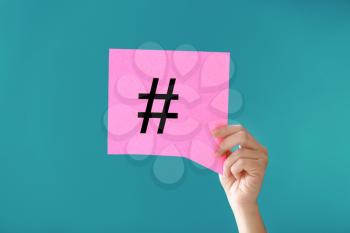 Female hand holding sheet of paper with hashtag sign on color background�