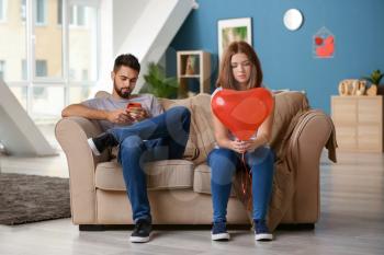 Sad young woman holding heart-shaped balloon near indifferent man playing with mobile phone at home. Relationship problems�