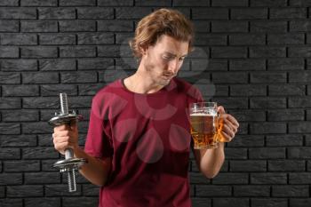 Young man with dumbbell and mug of beer near black brick wall. Concept of choice between alcohol and sport�