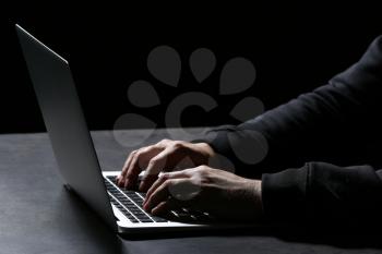 Professional hacker with laptop sitting at table on dark background, closeup�