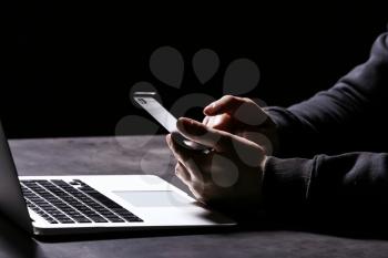 Professional hacker with laptop and mobile phone sitting at table on dark background, closeup�