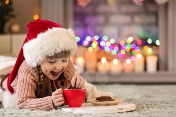 Cute little girl in Santa hat drinking hot chocolate and eating cookies at home on Christmas eve�