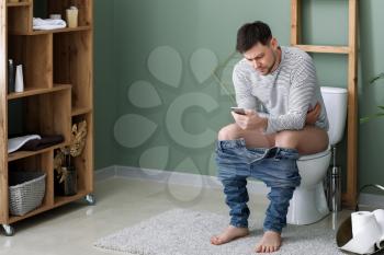 Man with mobile phone suffering from diarrhea while sitting on toilet bowl at home�