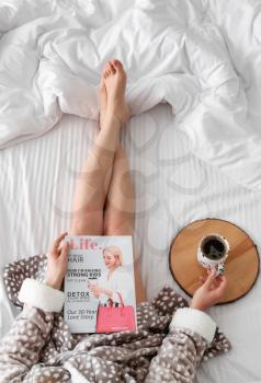 Young woman with fashion magazine drinking coffee on bed in morning�