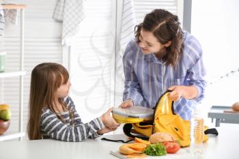 Mother with cute daughter packing lunch box into backpack at home�
