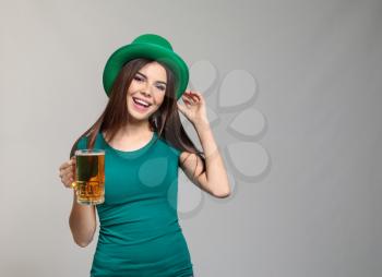 Beautiful young woman in green hat and with mug of beer on grey background. St. Patrick's Day celebration�