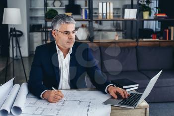 Mature architect working with laptop and drawings in office�