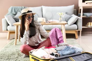 Young woman talking by phone while packing suitcase at home. Travel concept�