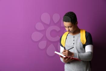 African-American schoolboy writing in notebook on color background�