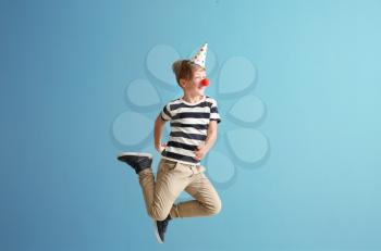 Jumping little boy with clown nose and party hat against color wall. April fools' day celebration�