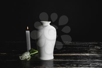 Mortuary urn with burning candle and flower on table against dark background�