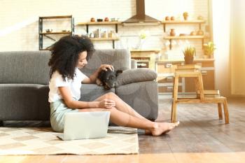 Beautiful African-American woman with cute funny dog working on laptop at home�