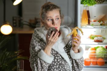 Beautiful young woman eating unhealthy food near refrigerator at night�