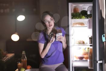Wary young woman eating unhealthy food near fridge in kitchen at night�