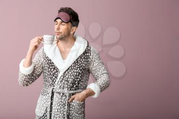 Funny young man with sleep mask and cup of coffee on color background�