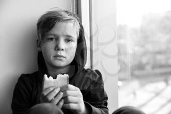 Homeless little boy with bread sitting on window sill indoors�