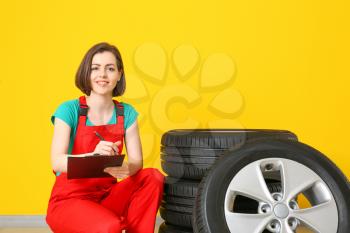 Young female mechanic in uniform and with car tires near color wall�