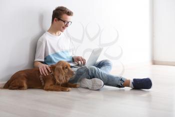 Teenage boy with cute dog and laptop sitting near light wall�