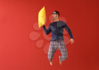 Jumping young man in pajamas with sleep mask and pillow on color background�