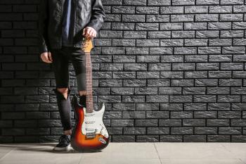 Handsome young man with guitar near dark brick wall�