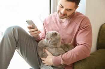 Young man with cute funny cat and mobile phone at home�